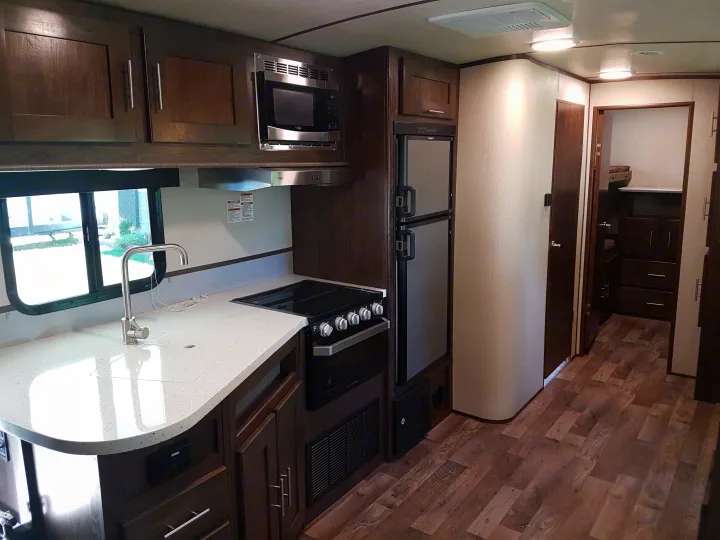 An RV rental's spacious kitchen area, with dark wood cabinets, light speckled countertops, built-in stove, microwave, and refrigerator.. Cruiser Rv Corp Radiance 2019