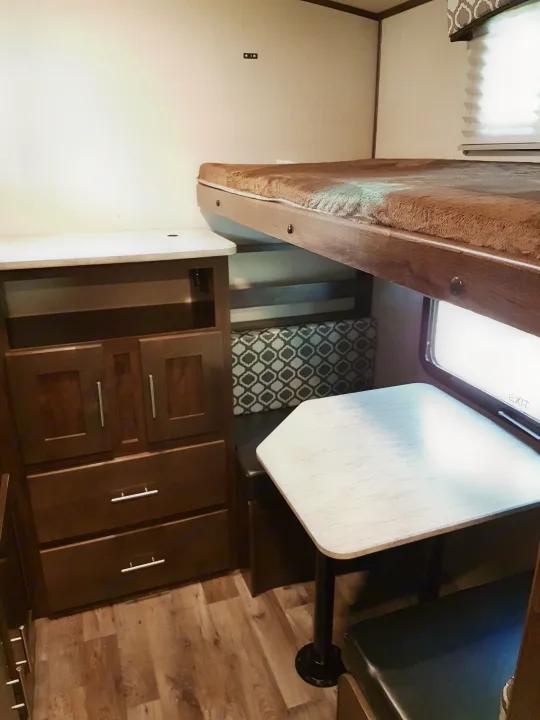 RV rental interior: dinette with patterned bench, light wood table, dark cabinets, wood-look floor, and upper bunk bed.. Cruiser Rv Corp Radiance 2019