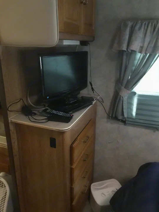 Cozy RV rental interior: A Sharp TV on a wooden dresser with Sony remotes, a window with curtains, and two fans.. Gulf Stream Sun Stream 1995