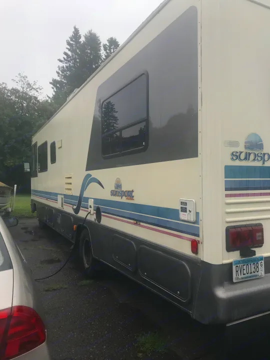 White Sun Sport RV rental (motorhome) parked on gravel, power cord connected. Features blue/purple stripes; MN plate RVE0138.. Gulf Stream Sun Stream 1995