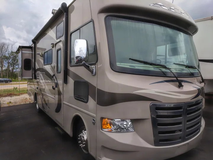 Modern beige RV rental with a slide-out extended, parked on asphalt under cloudy skies, ready for adventure.. Thor Motor Coach A.C.E 2015