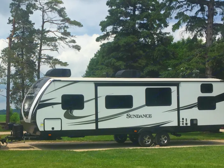 This Sundance Ultra-Lite travel trailer RV rental, white with black accents, is parked amidst trees for a relaxing camping trip.. Heartland Sundance 2018