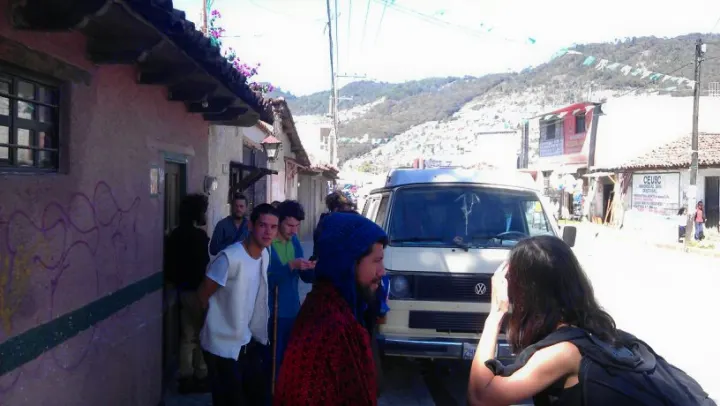 People gather around an RV rental (white VW van) on a sunny street in a hilly town, surrounded by buildings.. Volkswagen Vanagon 1984