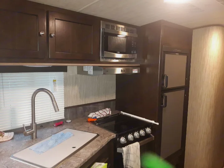 Well-equipped RV rental kitchen interior with dark wood cabinets, sink, microwave, stove, and refrigerator for travel comfort.. Heartland Sundance 2018