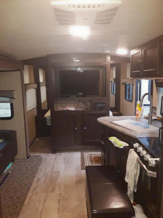 Cozy rv rental interior featuring a kitchen with dark cabinets, living area, and entertainment center with a TV. Light tiled floor.. Heartland Sundance 2018