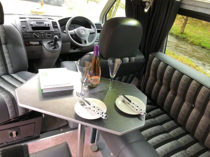 Cozy RV rental interior: dining setup with a river view. Features table, plates, drinks, book, and comfortable seats.. Volkswagen T5 Vivante 2015