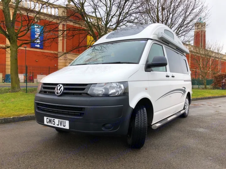 White VW camper van (GM15 JVU) for RV rental, parked on asphalt with a large brick building and bare trees in the background.. Volkswagen T5 Vivante 2015