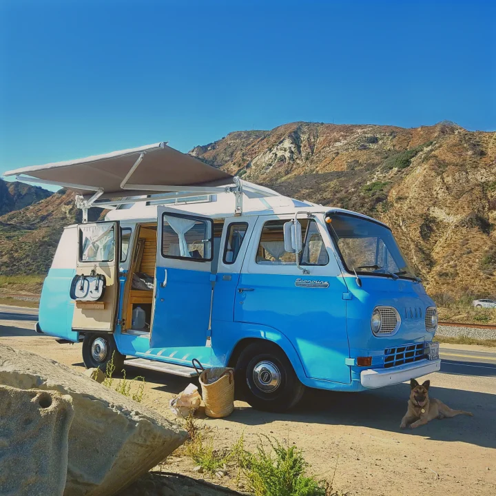 Blue & white vintage camper van (RV rental) under extended awning, mountains backdrop, dog resting. Ready for adventure!. Ford Econoline 1966