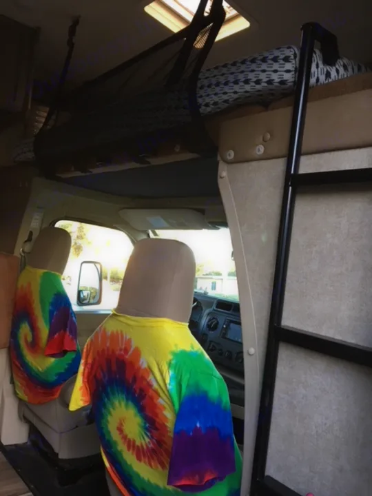 Inside a vibrant **rv rental**: tie-dye seat covers, an overhead bunk bed with ladder, and a handy storage net.. Coachmen Freelander 2016