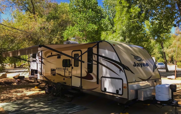 A tan Jayco White Hawk travel trailer with awning extended is parked at a sunny, tree-lined campsite. Perfect RV rental.. Jayco White Hawk Ultra Lite 2015