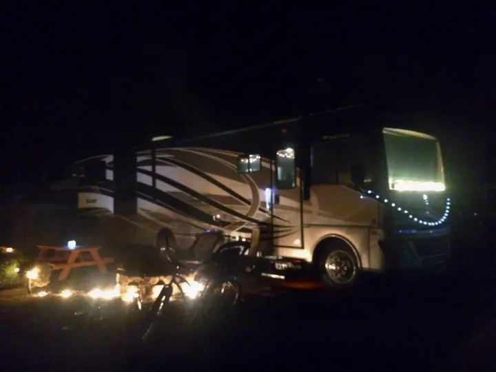Illuminated RV rental at a nighttime campsite, with a glowing fire pit, picnic table, and bike.. Fleetwood Bounder 2015