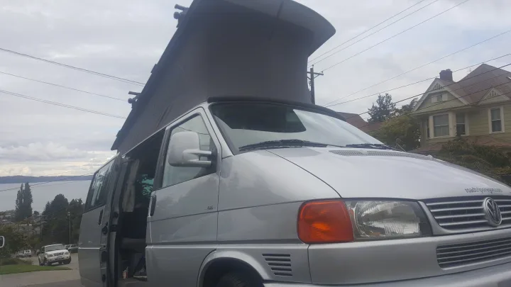 Silver pop-top camper van, ideal for RV rental. Parked with a water view, its side door open for adventure.. Volkswagen Westfalia 2002