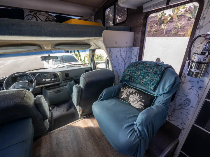 Cozy RV rental interior: driver's area, overhead bunk, and a comfy armchair. Features wood-look floor & scenic wall decor.. Other Other 1992