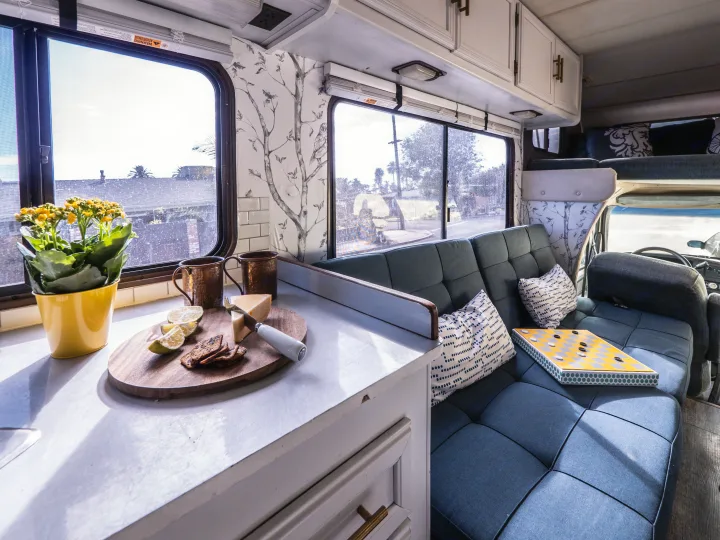 Inside a modern RV rental: counter with cheese, a cozy blue sofa, and nature-inspired wallpaper create a comfy travel space.. Other Other 1992