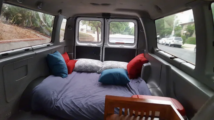 Cozy RV rental interior with a full bed & pillows. Views out side & back windows of parked cars & trees.. Ford E350 2000