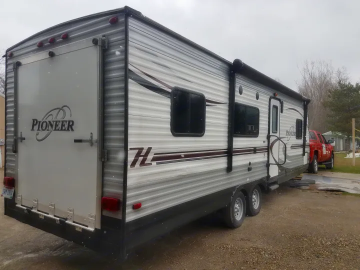 Grey Pioneer travel trailer RV rental with a rear ramp, parked on gravel. A red truck is seen behind it on a cloudy day.. Heartland Pioneer 2016