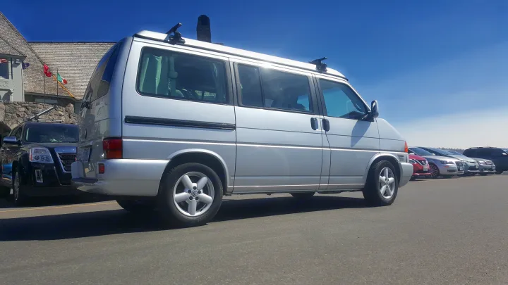 Silver VW T4 camper van, ideal for an RV rental vacation, parked in a sunny lot near a building with flags.. Volkswagen Westfalia 2002