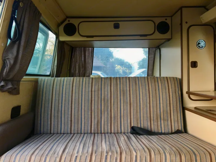 Interior view of an RV rental: striped bench seating that converts to a bed, overhead cabinets, and rear windows with curtains.. Volkswagen Vanagon 1984