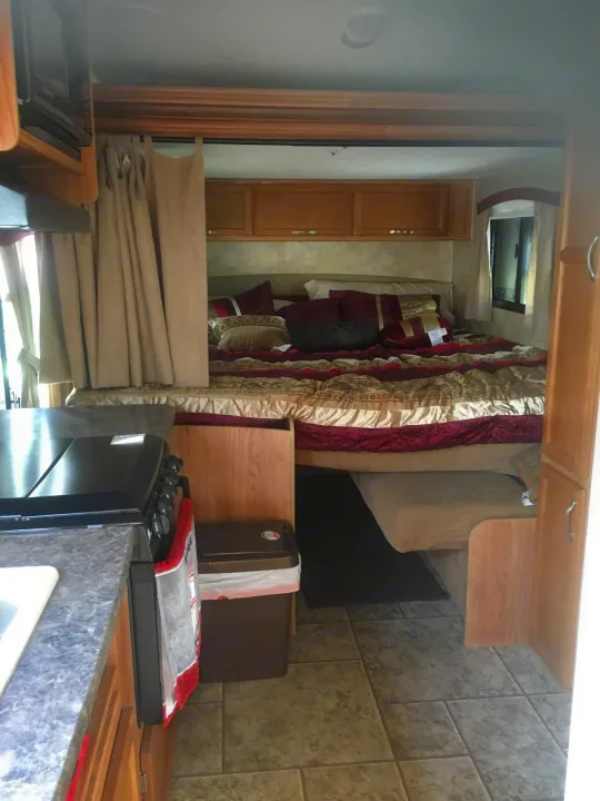 View inside an RV rental showing a full bed with patterned bedding, overhead cabinets, and a side window. Kitchen on left.. Jayco Jay Feather Ultra Lite 2012