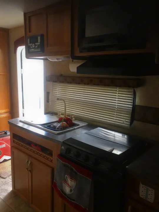 Explore this functional RV rental kitchen: wooden cabinets, sink, microwave, and stove with a cozy coffee towel. Light streams from an open door.. Jayco Jay Feather Ultra Lite 2012