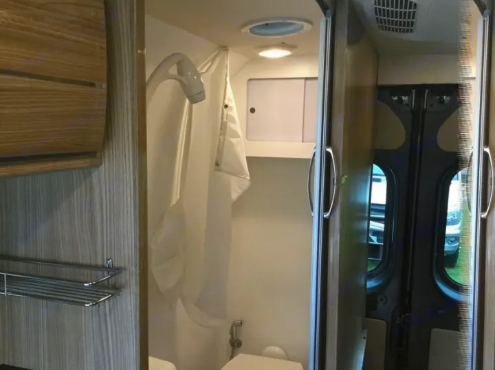 RV rental interior: a compact bathroom with shower, toilet, and shelving, looking towards the rear doors of the van.. Winnebago Travato 2019