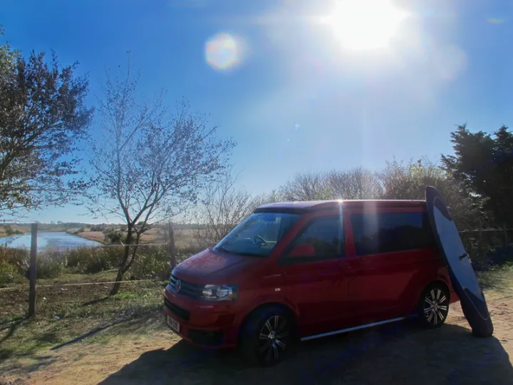 Sunny day with a red camper van and paddleboard by a scenic river. Ideal RV rental for water sports getaways.. Volkswagen T5 premium camper 2011