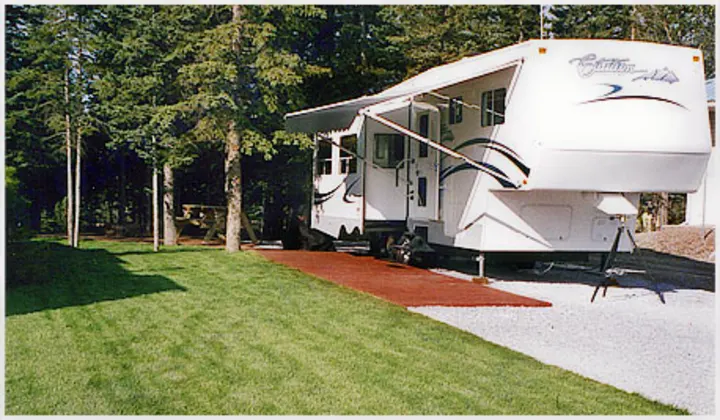 White fifth-wheel rv rental parked on a gravel site, with an extended awning, green grass, and tall pine trees in the background.. Citation Supreme 29CKS 2012
