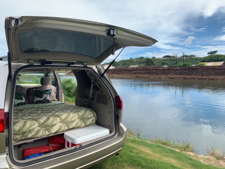Cozy minivan RV rental setup by a calm river. Features a bed with tropical bedding & cooler, perfect for van life adventures.. Toyota Sienna Camper 2004