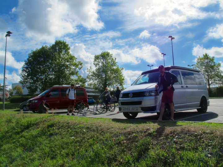 Friends by a silver RV rental van & red van with bikes in a sunny parking lot.. Volkswagen T5 premium camper 2012