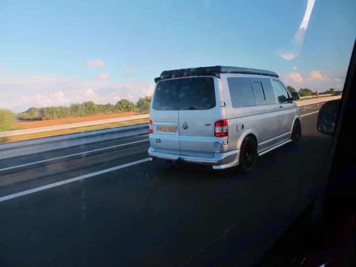 A silver VW camper van, potentially an rv rental, drives on a highway with green trees and blue sky in the background.. Volkswagen T5 premium camper 2012