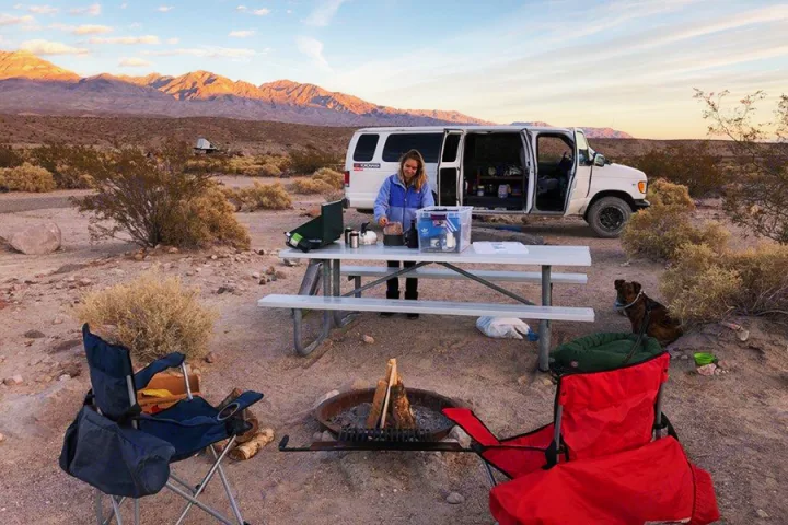 RV rental desert camping: Woman cooking at camp beside her van with a dog & campfire, mountains under sunset glow.. Ford E350 2000