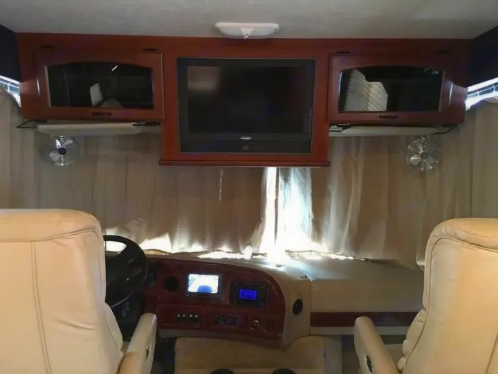 Inside an RV rental: front cabin with cream seats, dashboard, TV, and side fans above windshield curtains.. Ford Hurricane 2011