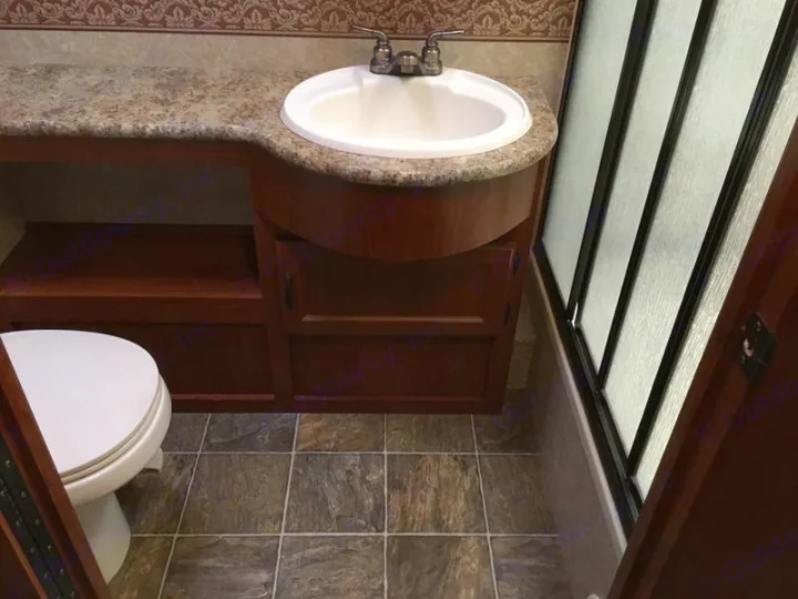 Compact RV rental bathroom: dark wooden vanity, light speckled countertop with sink, toilet, tile floor, and frosted shower.. Ford Hurricane 2011