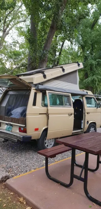 A tan pop-top camper van (RV rental) is set up for camping at a shaded site with a picnic table.. Volkswagen Westfalia 1982