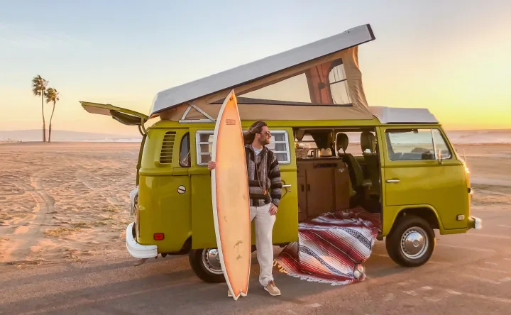Sunset beach vibes in a vintage VW RV rental. A man with a surfboard stands beside his pop-top camper van by the ocean.. Volkswagen Westfalia 1977