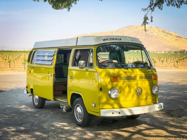 Bright yellow VW Westfalia camper van for RV rental, parked in a sunny vineyard with mountains, ideal for nature travel.. Volkswagen Westfalia 1977