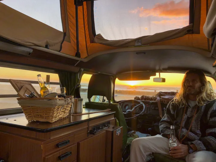 Scenic 'rv rental' view: Man relaxing in a camper van, enjoying a drink as the sun sets over the crashing ocean waves.. Volkswagen Westfalia 1977