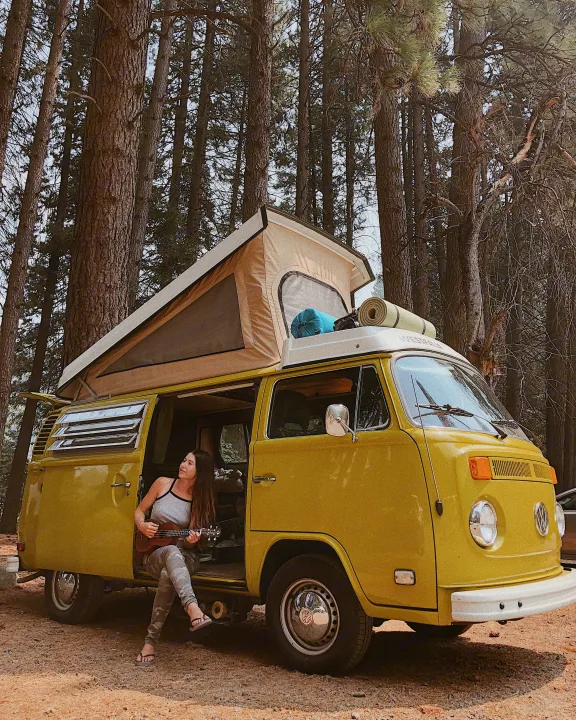 RV rental ready: A woman plays ukulele in her classic yellow VW camper van with a pop-top roof, parked in a serene forest.. Volkswagen Westfalia 1977