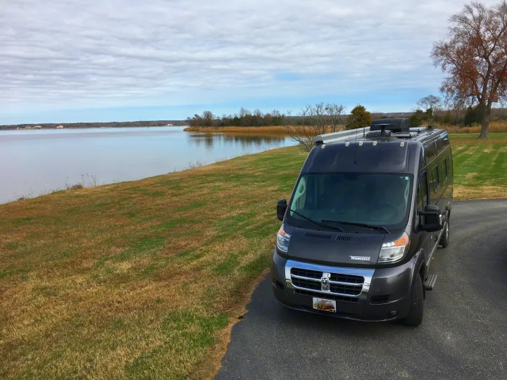 A grey Winnebago RV van is parked on asphalt beside a grassy slope leading to a calm bay under a cloudy sky, ideal for an RV rental.. Winnebago Travato 59K 2019