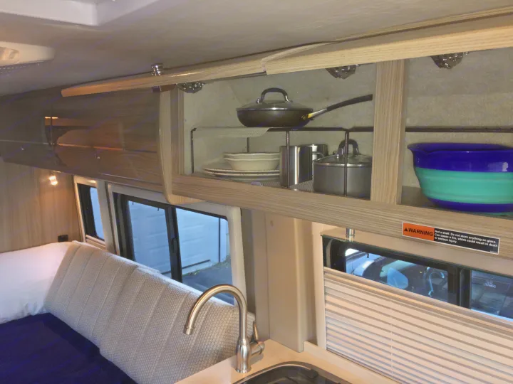 Inside an RV rental, showing the kitchen area with sink, full cabinets of cookware/dishes, and a bed. A warning label about glass/fire is visible.. Winnebago Travato 59K 2019