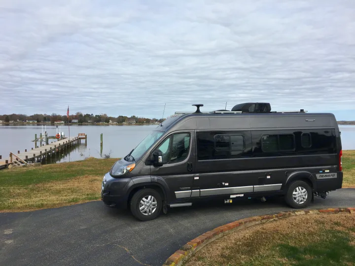 This dark gray camper van, a potential RV rental, is parked beside a wooden pier extending into a calm bay. Overcast sky.. Winnebago Travato 59K 2019