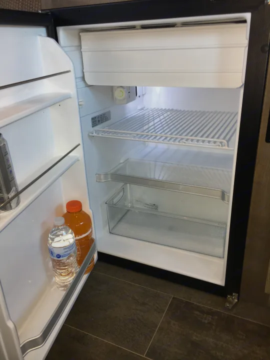 Inside an RV rental: a clean, open mini-fridge with its shelves, crisper drawer, and water/juice bottles in the door.. Winnebago Travato 59K 2019