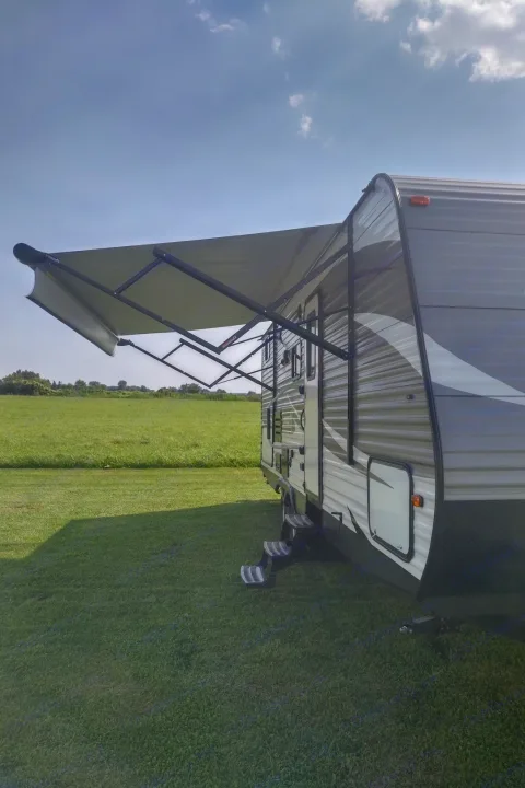 An RV rental, a grey travel trailer with an extended awning, sits in a sunny green field, perfect for outdoor adventures.. Coleman 244BH 2017
