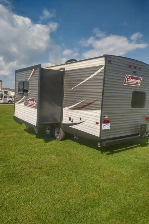 This Coleman RV rental, a gray travel trailer with its slide-out extended, is parked on a vibrant green field.. Coleman 244BH 2017