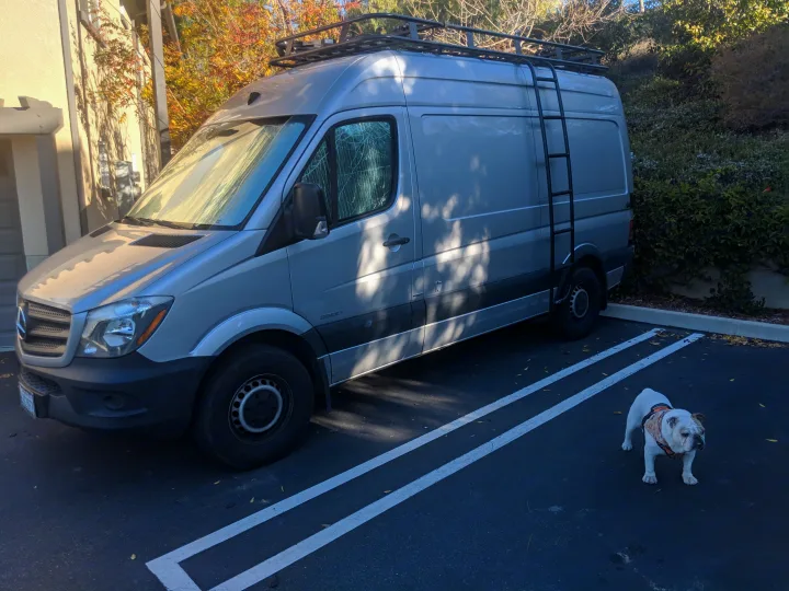 A silver Mercedes Sprinter van, equipped as an RV rental with a roof rack and ladder, parked outdoors. A white bulldog stands in the foreground.. Mercedes-Benz Sprinter 2016