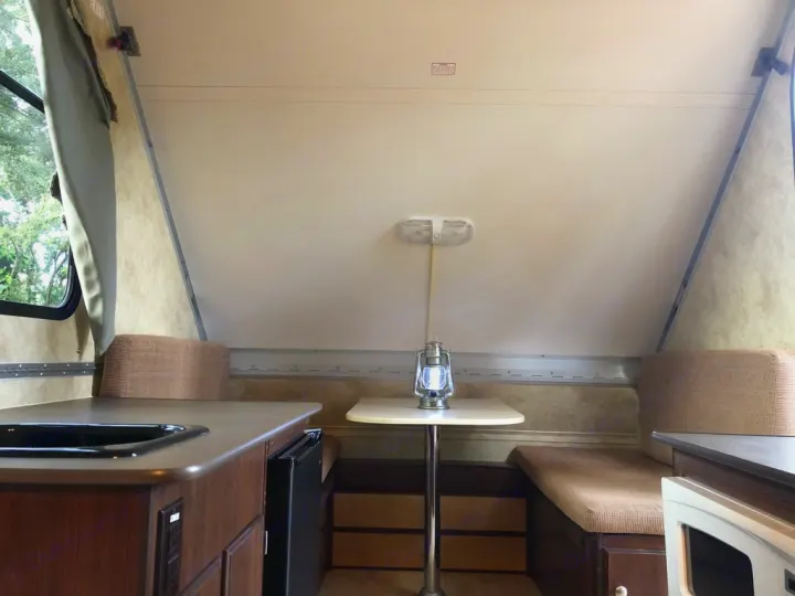 Interior of an RV rental: a compact living space with a dining table, padded benches, and kitchen counter with a sink, next to a window.. A-Liner Scout 2013