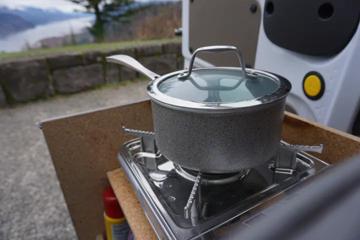 A speckled gray pot with a glass lid sits on a portable gas stove, part of an RV rental kitchen setup with a scenic outdoor view.. Ford Transit Custom 2017