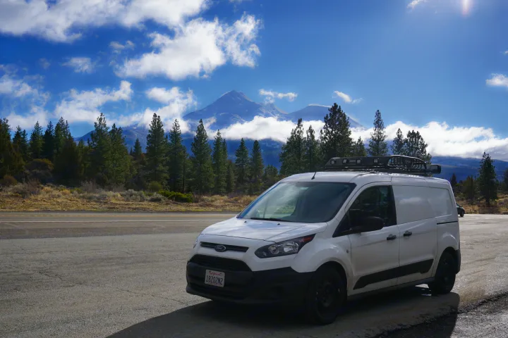 White camper van (RV rental) parked by a road with a majestic snow-capped mountain & pine trees. Perfect for adventure!. Ford Transit Custom 2017