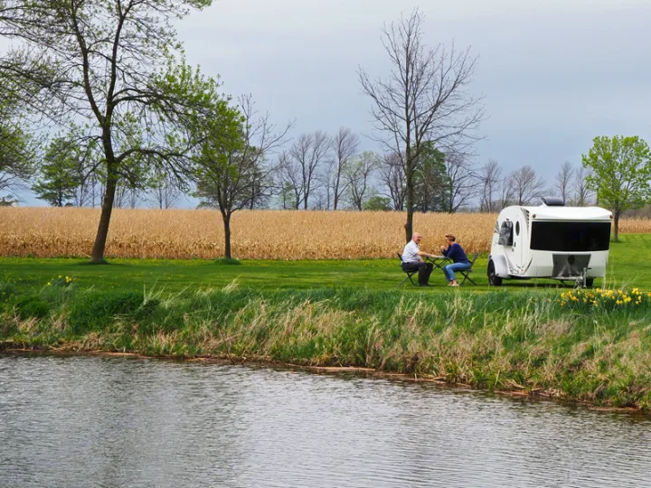 A couple enjoys an RV rental by a pond, with a white camper and golden field visible in this serene outdoor scene.. InTech RV Luna 2018