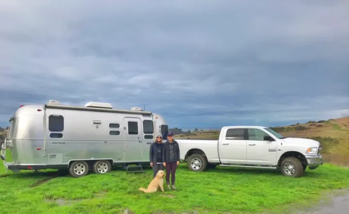 Two people and their dog stand beside a silver Airstream RV rental and a white pickup truck on a grassy hill.. Airstream Safari 2008